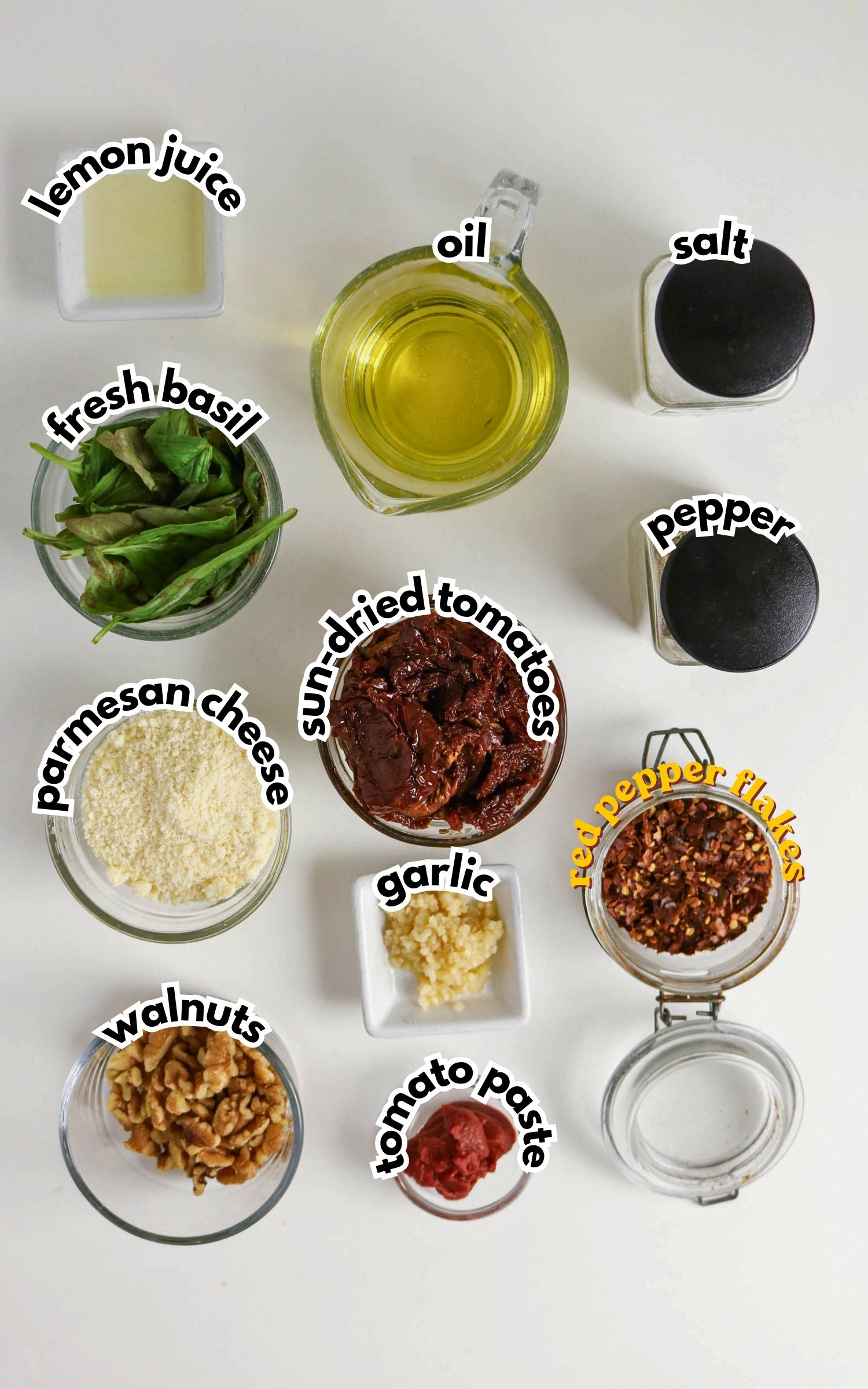 Top-down view of ingredients for a recipe, including lemon juice, oil, salt, pepper, fresh basil, parmesan cheese, sun-dried tomatoes, garlic, red pepper flakes, walnuts, and tomato paste.
