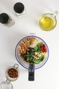 A food processor filled with basil, walnuts, sun-dried tomatoes, parmesan, and garlic, surrounded by oil, spices, and red pepper flakes on a white surface.