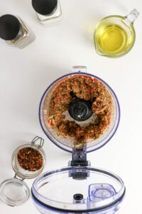 A food processor with a blended herb and spice mixture, surrounded by oil, spices, and red pepper flakes on a white countertop.