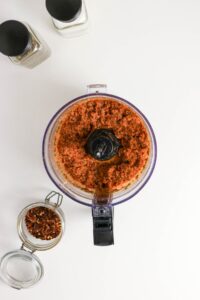 A food processor filled with red salsa sits on a white surface, next to a jar of red pepper flakes and salt and pepper shakers.