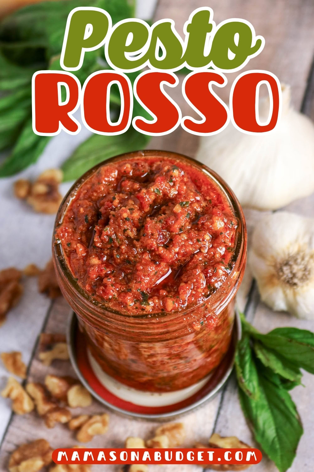 A jar of pesto rosso surrounded by fresh basil leaves, walnuts, and garlic, with Pesto Rosso text above and mamasanabudget.com at the bottom.