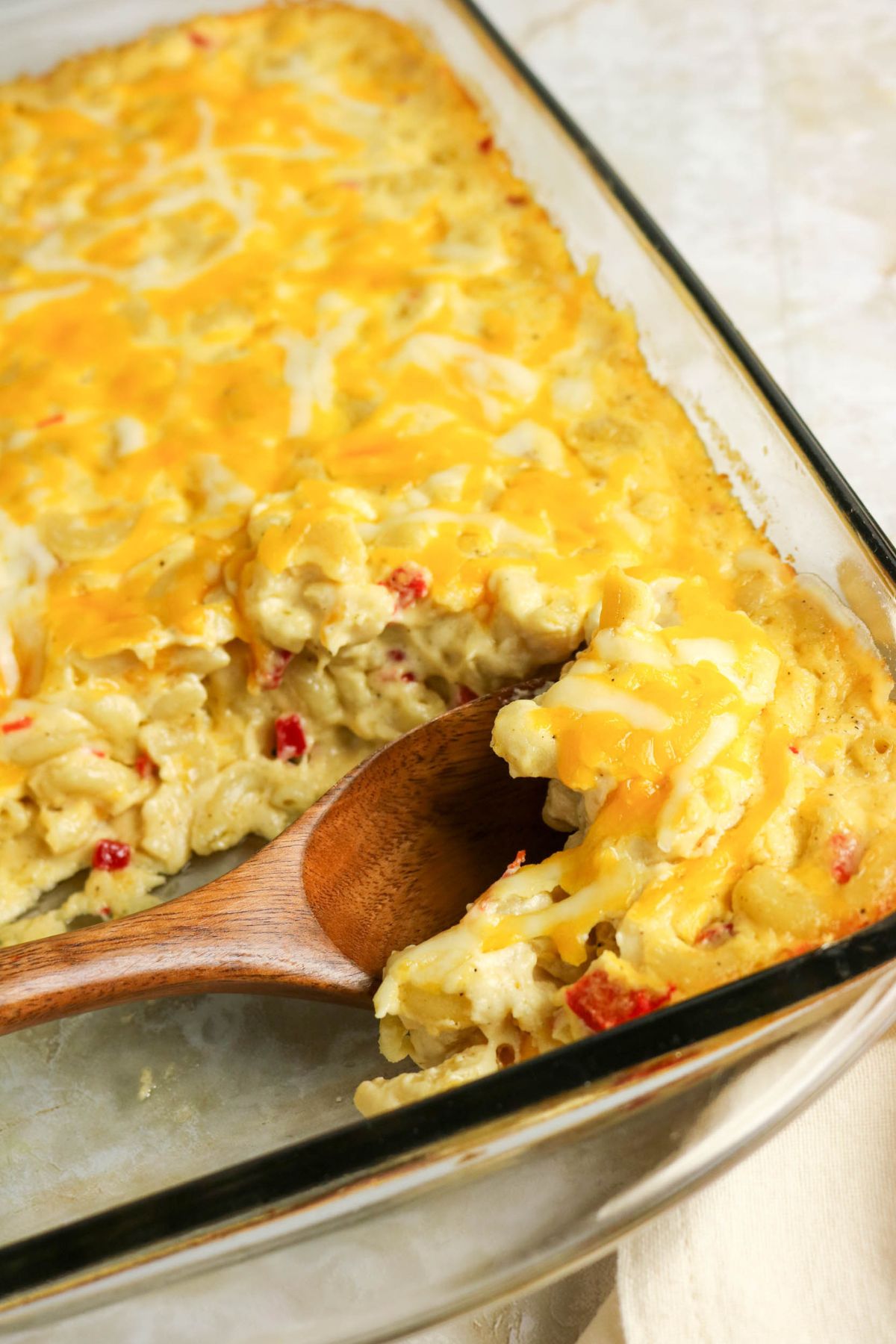 A glass baking dish with baked macaroni and cheese topped with melted cheese. A wooden spoon is lifting a serving from the dish.