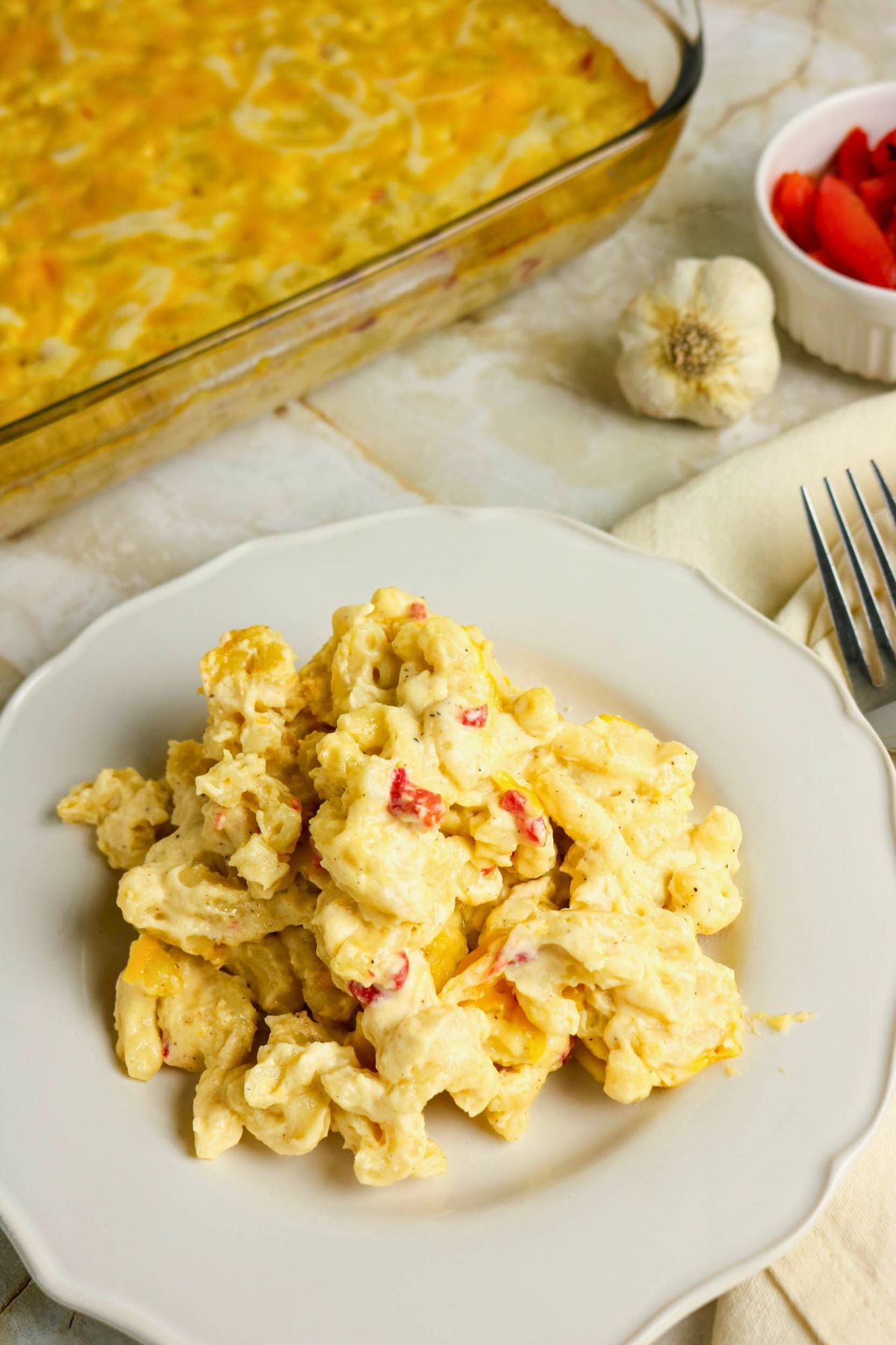A serving of cheesy scrambled eggs with red peppers on a white plate, with a casserole dish, garlic bulb, and a bowl of sliced strawberries in the background.