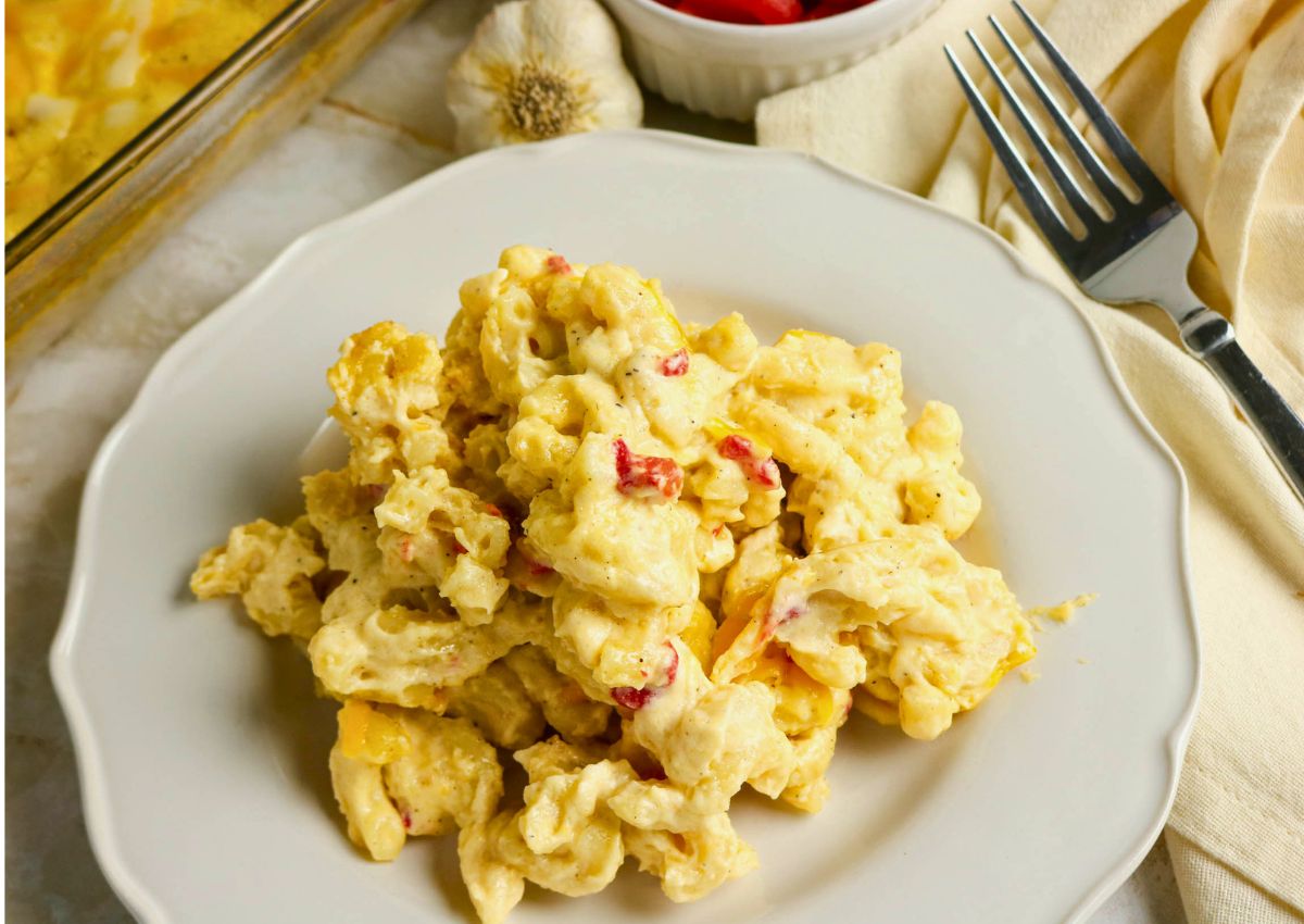 A serving of creamy macaroni and cheese with pimentos on a white plate, next to a fork, napkin, and a bowl of sliced red peppers.