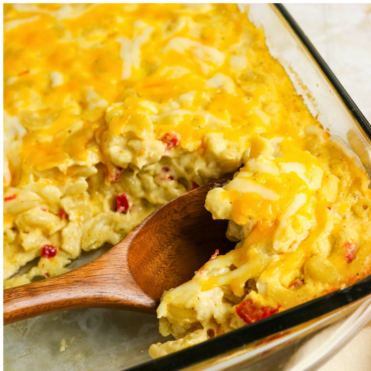 A wooden spoon serves a portion of cheesy macaroni casserole with visible pieces of red pepper from a glass baking dish.