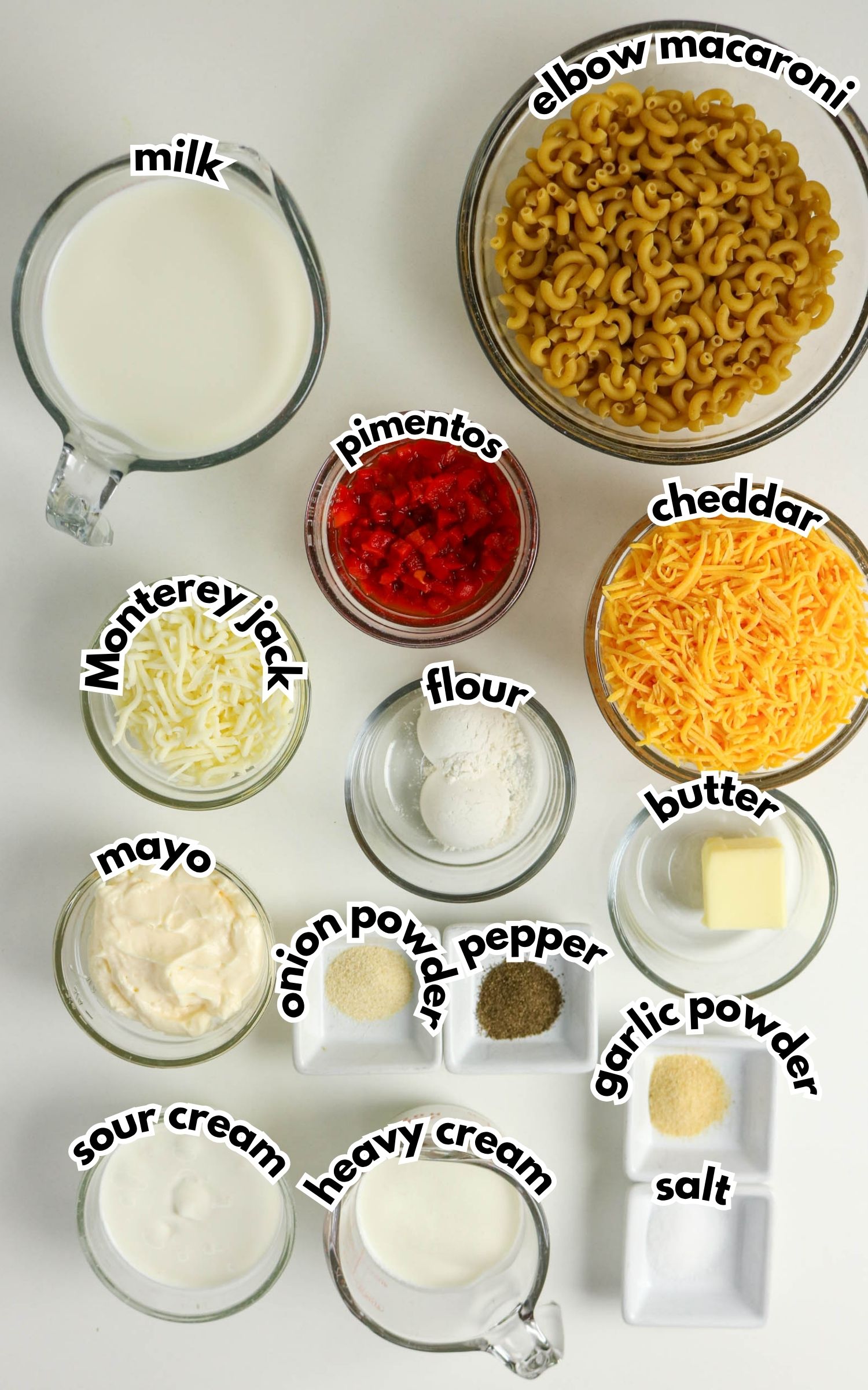 Ingredients for macaroni and cheese are arranged in bowls and cups, including macaroni, cheeses, milk, cream, butter, flour, seasonings, mayo, sour cream, and pimentos.