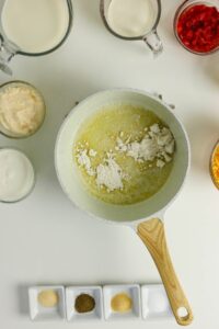 A saucepan with melted butter and flour is surrounded by ingredients including milk, cream, cheese, seasonings, and diced peppers, arranged on a white surface.