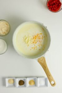 A white saucepan with milk and shredded cheese sits on a white surface, surrounded by small bowls of spices, chopped red peppers, and other ingredients.