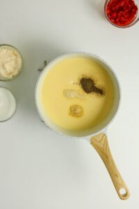 A white saucepan with a creamy yellow mixture and various seasonings on top sits on a white surface, surrounded by small bowls of mayonnaise, milk, and chopped red peppers.