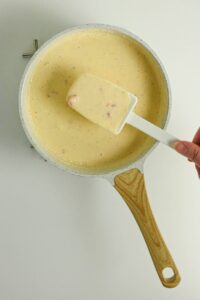 A hand holds a white spatula stirring a creamy yellow sauce with small chunks in a white pan on a light surface.