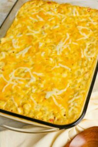 A baked casserole topped with melted cheese in a rectangular glass dish, resting on a light-colored cloth with a wooden spoon nearby.