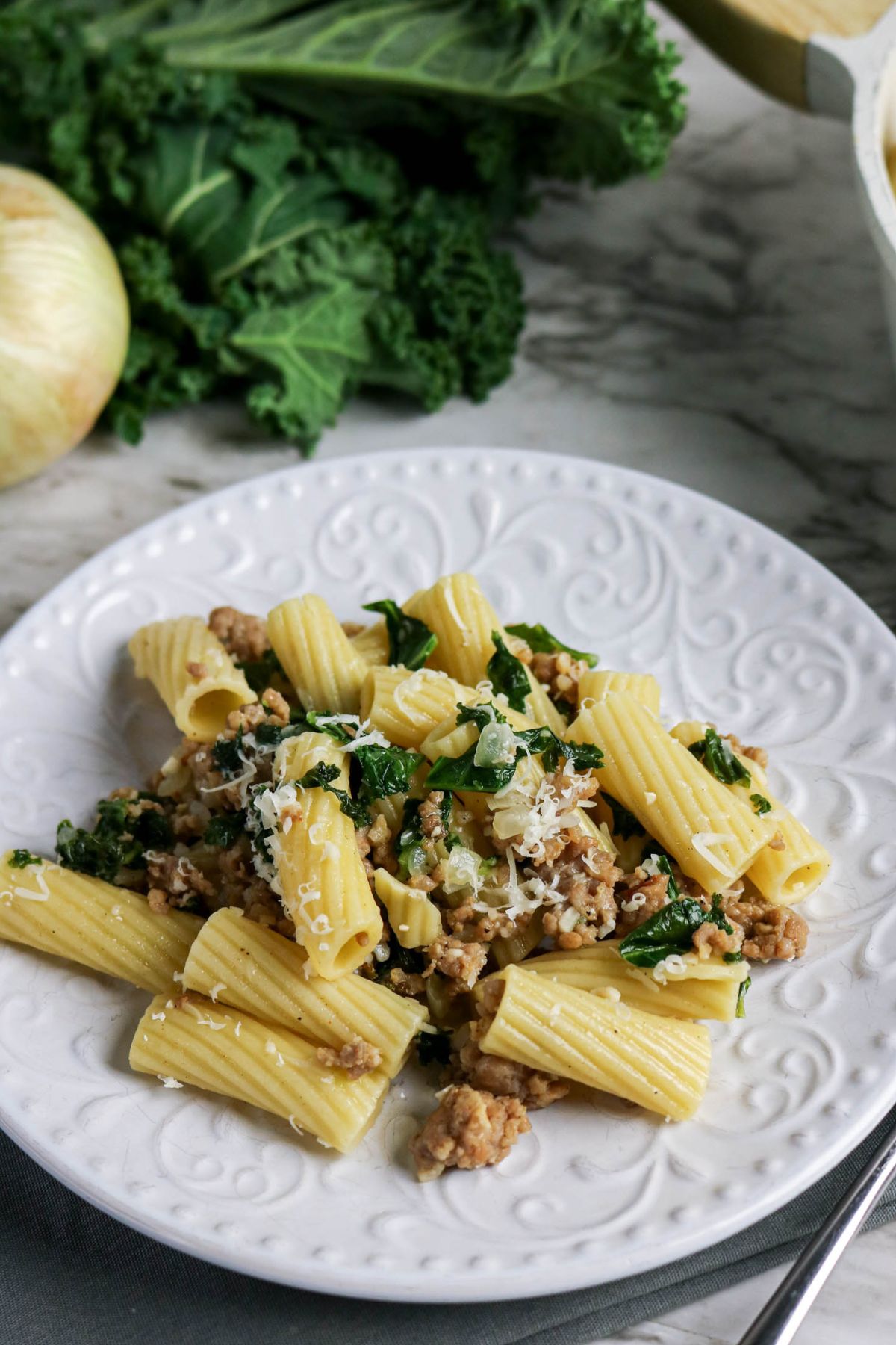 A white plate of rigatoni pasta with sausage, kale, and grated cheese, placed on a marble surface with a leafy green and onion in the background.