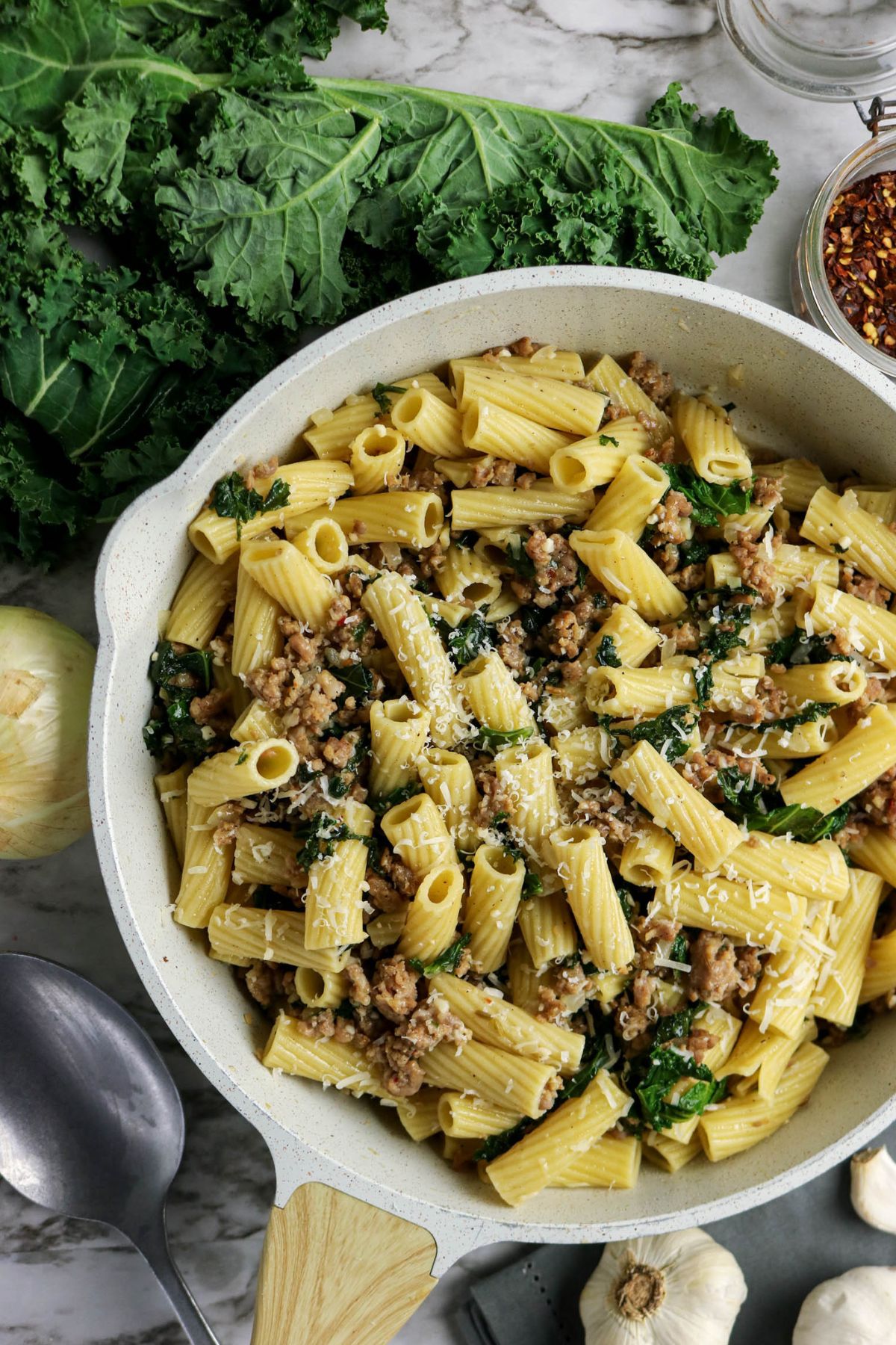 A skillet filled with rigatoni pasta, cooked ground meat, kale, and grated cheese, with fresh kale, onion, garlic, and red pepper flakes nearby.