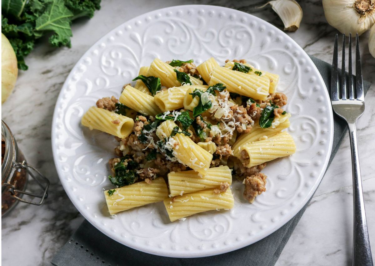 A white plate holds rigatoni pasta mixed with ground meat, kale, and grated cheese, set on a marble surface beside garlic, an onion, and a fork.
