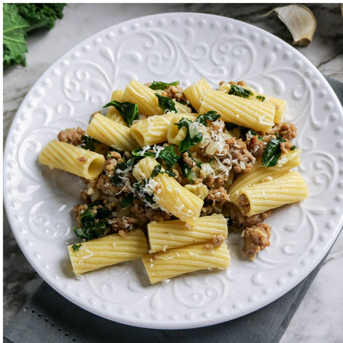 A white plate of rigatoni pasta with crumbled sausage, chopped kale, and grated cheese on a marble surface.