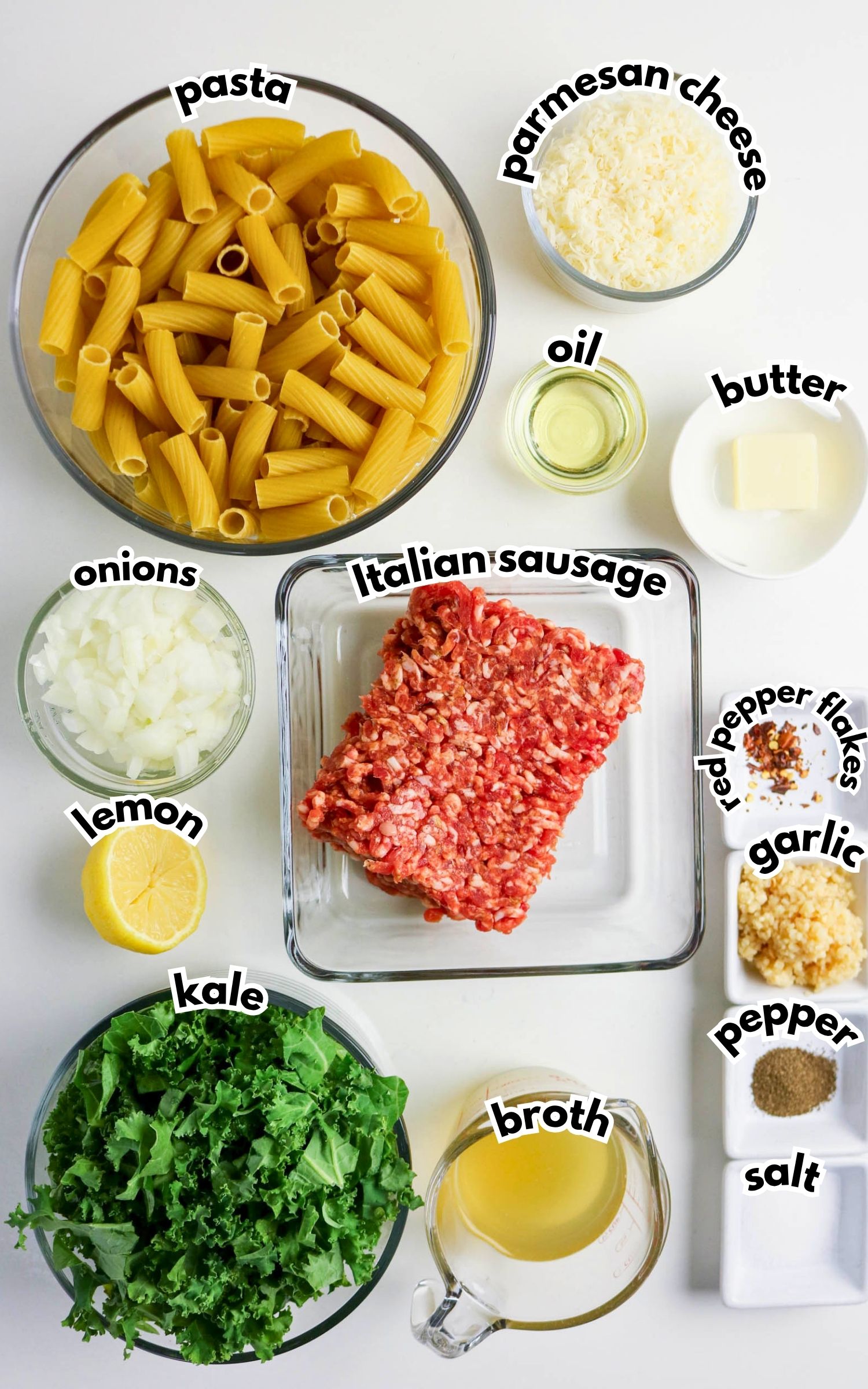 Top-down view of ingredients for a pasta dish, including rigatoni, Italian sausage, kale, onion, lemon, garlic, broth, parmesan, oil, butter, red pepper flakes, salt, and pepper.