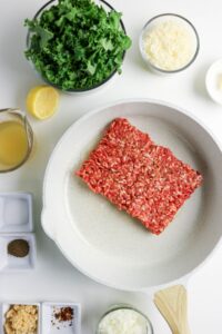 Raw ground meat in a white skillet surrounded by bowls of chopped kale, grated cheese, minced garlic, diced onion, spices, lemon, and broth on a white surface.