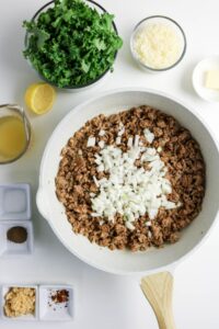 A skillet with cooked ground meat and chopped onions, surrounded by bowls of chopped kale, shredded cheese, broth, butter, lemon, spices, minced garlic, and red pepper flakes.