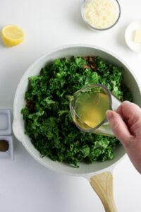 A hand pours broth from a glass measuring cup into a skillet filled with chopped kale. Nearby are a lemon half, a bowl of grated cheese, a pat of butter, and small dishes of spices.