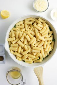 Large white skillet filled with uncooked rigatoni pasta on a white surface, surrounded by grated cheese, lemon half, butter, broth in a measuring cup, and small bowls of salt and pepper.