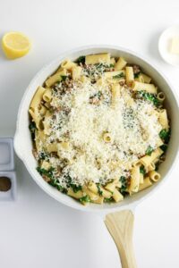 A white skillet filled with rigatoni pasta, leafy greens, and shredded cheese, next to a lemon wedge, a pat of butter, and small containers of pepper and salt on a white surface.