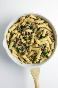 A white pan filled with rigatoni pasta, ground meat, and wilted greens, photographed from above on a white background.
