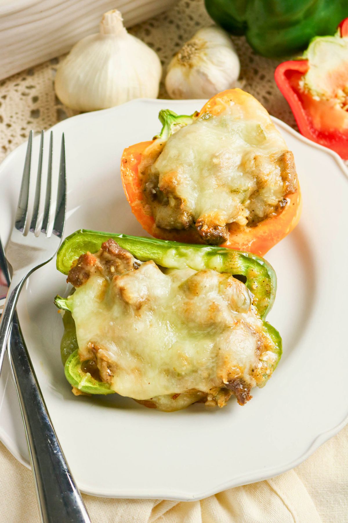A plate with two stuffed bell pepper halves topped with melted cheese, served with a fork and knife. Garlic bulbs and fresh peppers are in the background.