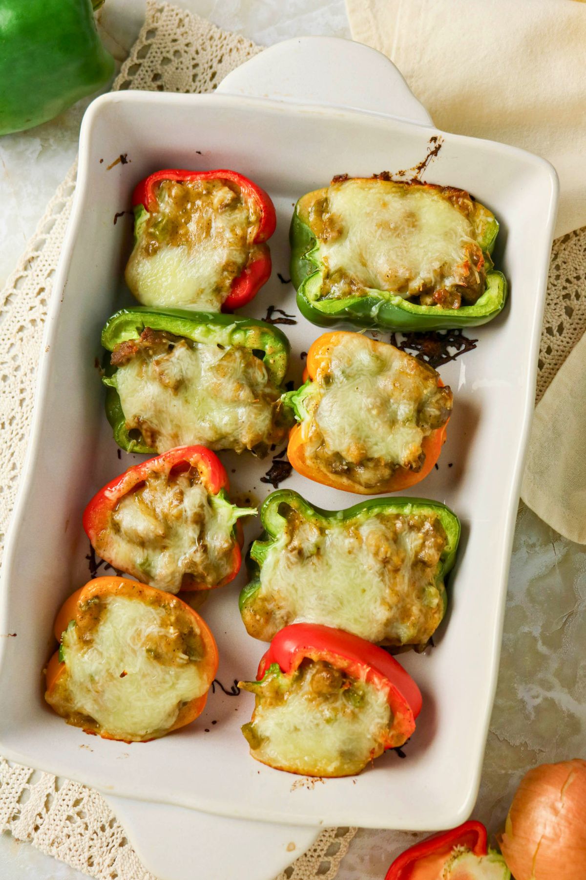 A white baking dish with eight stuffed bell pepper halves, topped with melted cheese, resting on a lace placemat.