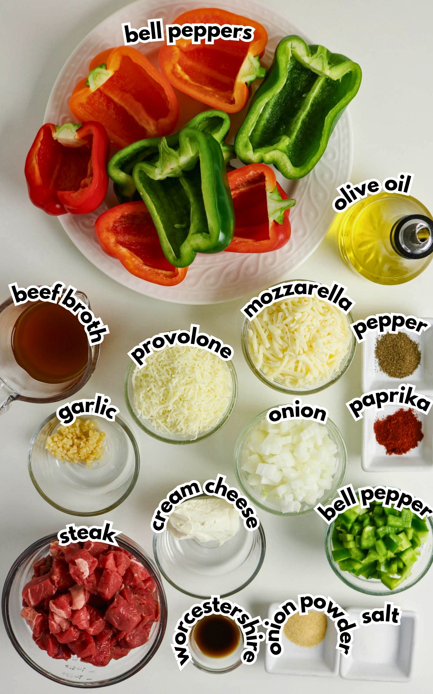 Overhead view of ingredients for a recipe, including chopped steak, bell peppers, cheese, onion, garlic, cream cheese, seasonings, beef broth, and olive oil arranged on a white surface.