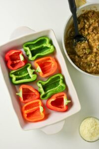 Halved red and green bell peppers in a baking dish beside a pan of cooked meat filling and a small bowl of shredded cheese.
