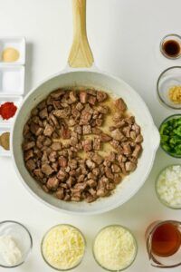 A white skillet with cooked diced beef surrounded by small bowls of chopped vegetables, shredded cheese, spices, and sauce ingredients on a white surface.