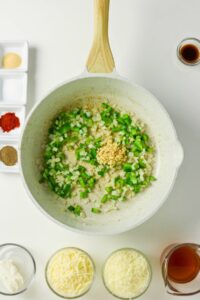 A white pan on a stovetop contains chopped onions, green bell peppers, and minced garlic, surrounded by small bowls of spices, shredded cheese, cream cheese, and liquids.