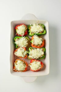 A white baking dish containing eight halved red and green bell peppers filled with stuffing and topped with shredded cheese, arranged in two rows.