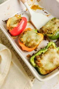 Four stuffed bell peppers with melted cheese on top are arranged in a white baking dish next to a spatula.