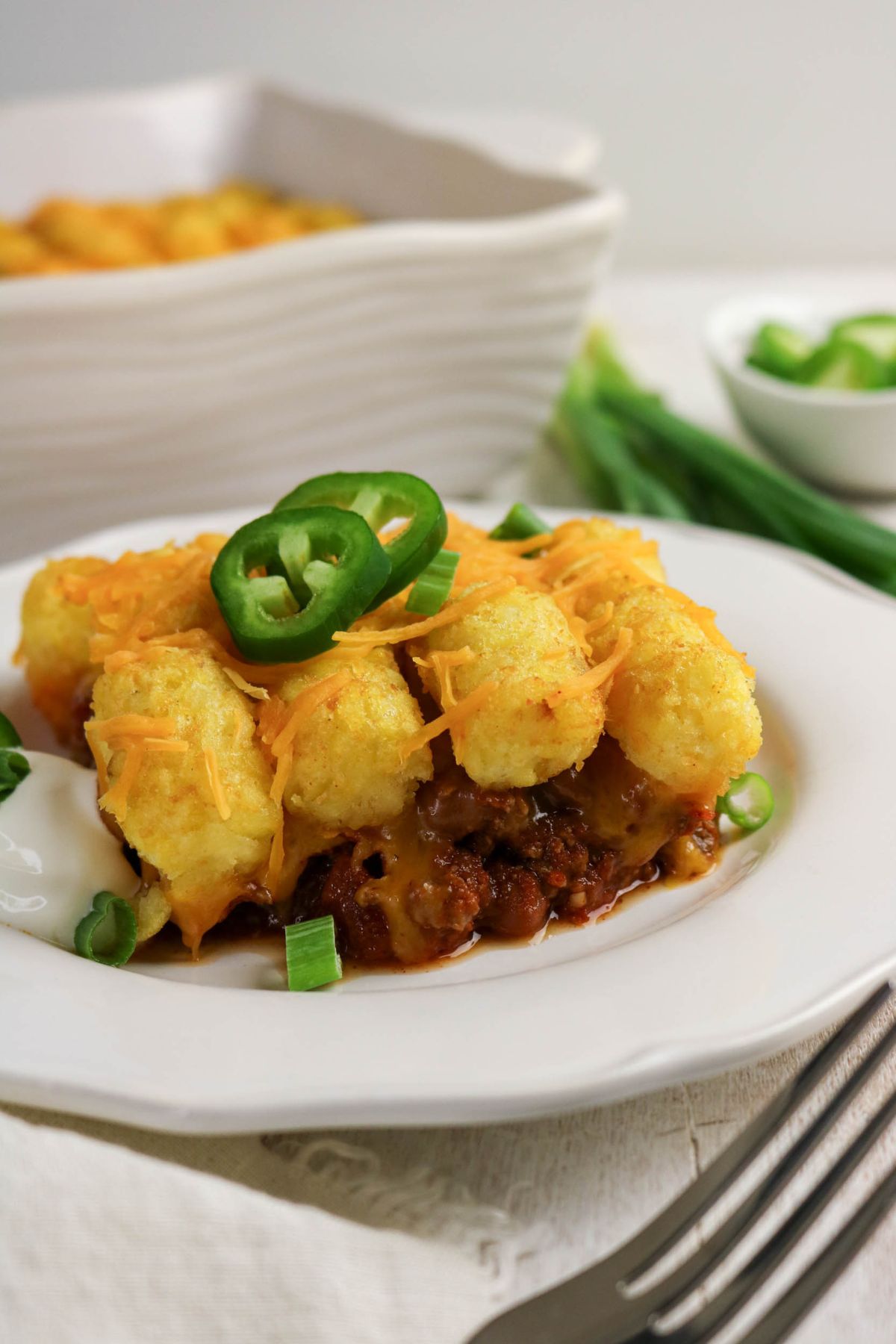 A serving of tater tot casserole topped with shredded cheese and sliced jalapeños, garnished with chopped green onions on a white plate.