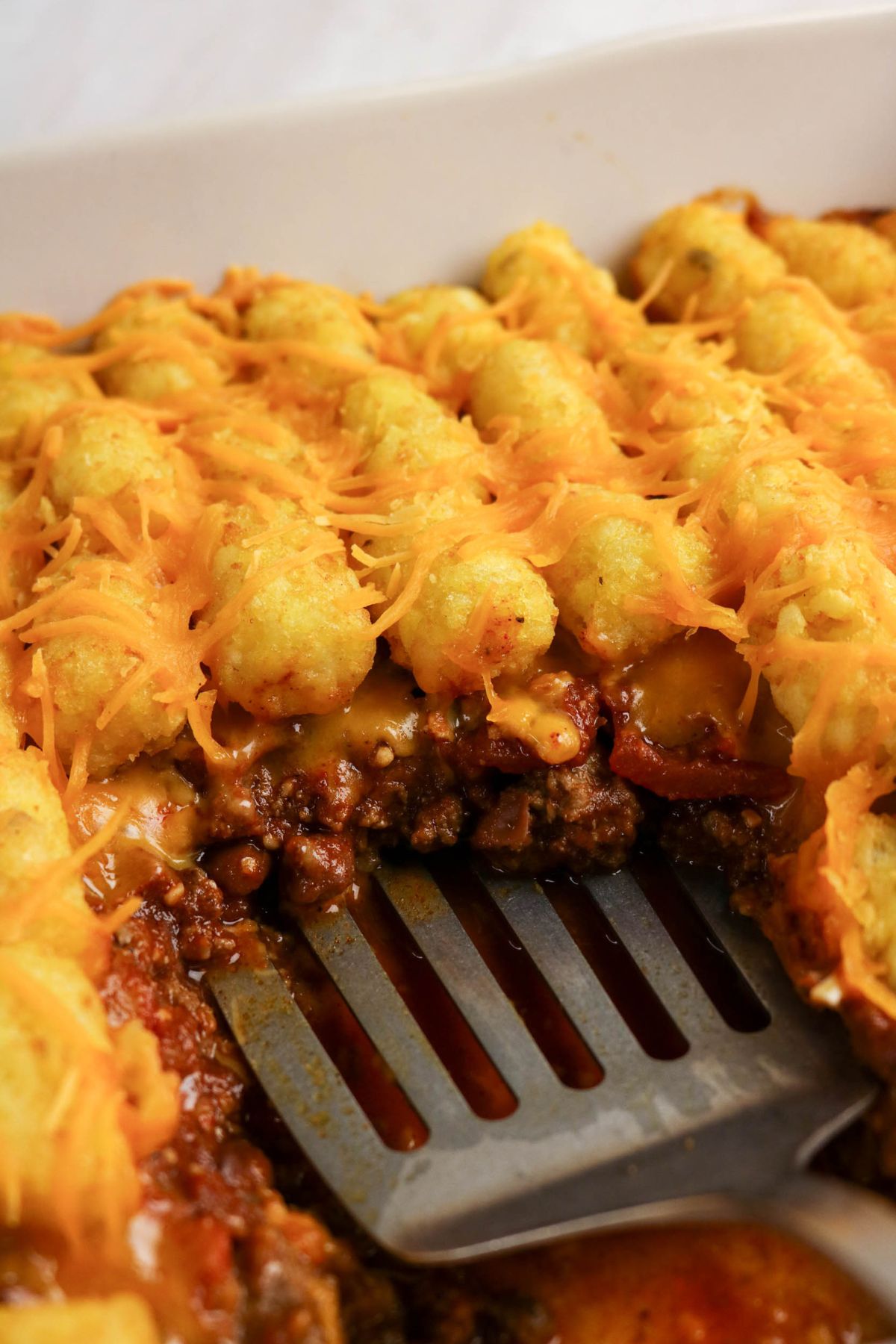 A close-up of a tater tot casserole in a white baking dish, showing layers of ground beef, sauce, cheese, and tater tots, with a spatula lifting a portion.