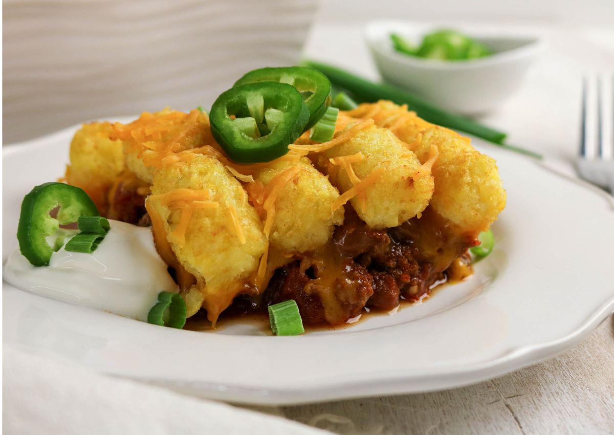 A plate of tater tot casserole topped with shredded cheese, sliced jalapeños, and green onions, with a dollop of sour cream on the side.