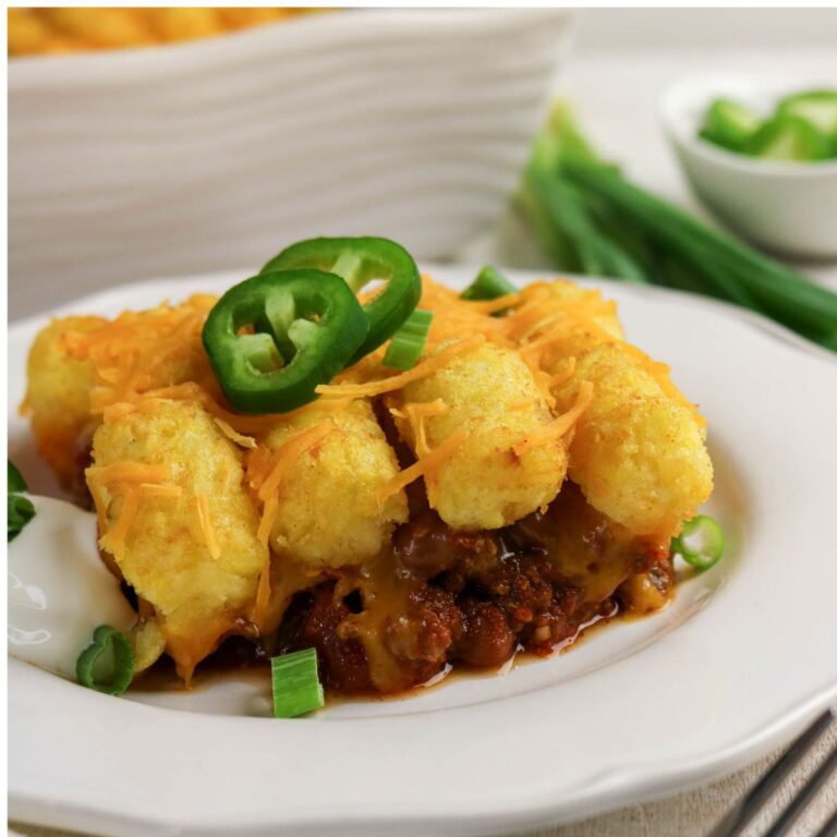 A serving of tater tot casserole topped with shredded cheese, sliced jalapeños, and green onions on a white plate.