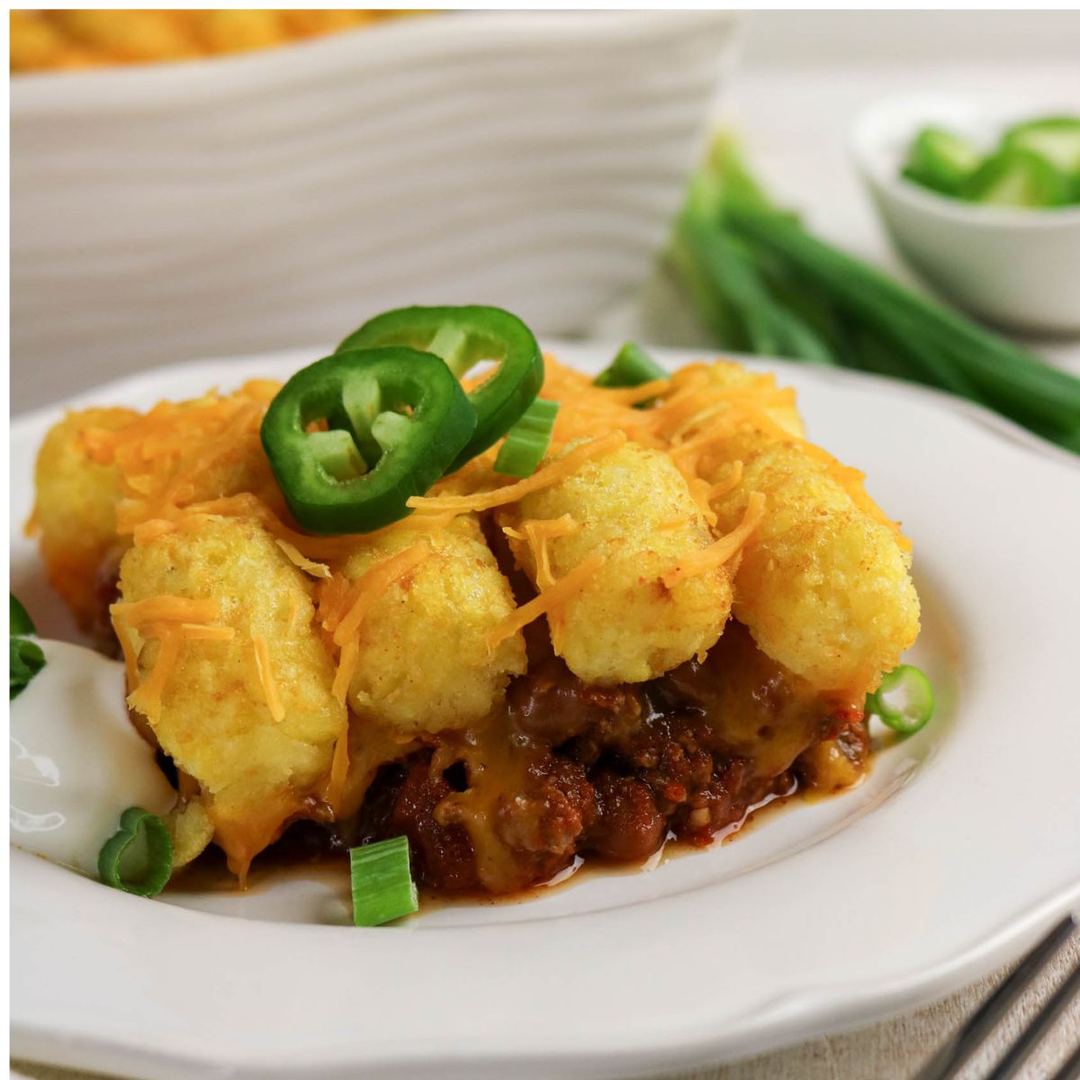 A serving of tater tot casserole topped with shredded cheese, sliced jalapeños, and green onions on a white plate.
