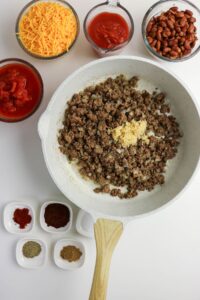 A skillet with cooked ground beef and minced garlic, surrounded by bowls of shredded cheese, beans, tomato sauce, diced tomatoes, and small dishes of various spices.