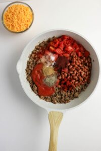 A skillet with browned ground beef, pinto beans, diced tomatoes, tomato sauce, and spices next to a bowl of shredded cheddar cheese.
