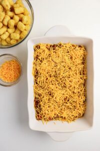A white baking dish filled with a layer of shredded cheddar cheese, next to bowls of frozen tater tots and more shredded cheese on a white surface.