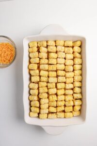 A white baking dish filled with neatly arranged frozen tater tots next to a small bowl of shredded cheddar cheese on a white surface.