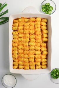 A rectangular casserole dish filled with tater tot casserole topped with melted cheese, surrounded by bowls of sour cream, sliced green onions, and sliced jalapeños.