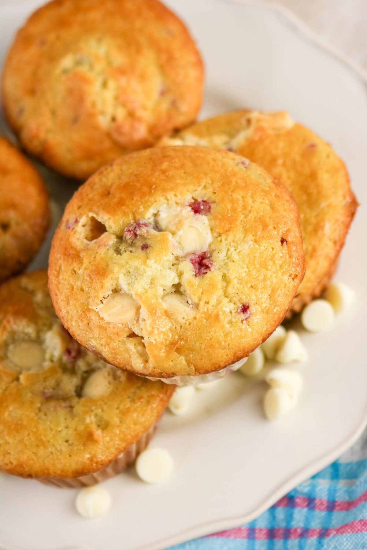 A plate of golden-brown muffins with visible white chocolate chips and berry pieces, arranged on a white plate next to a blue checkered cloth.