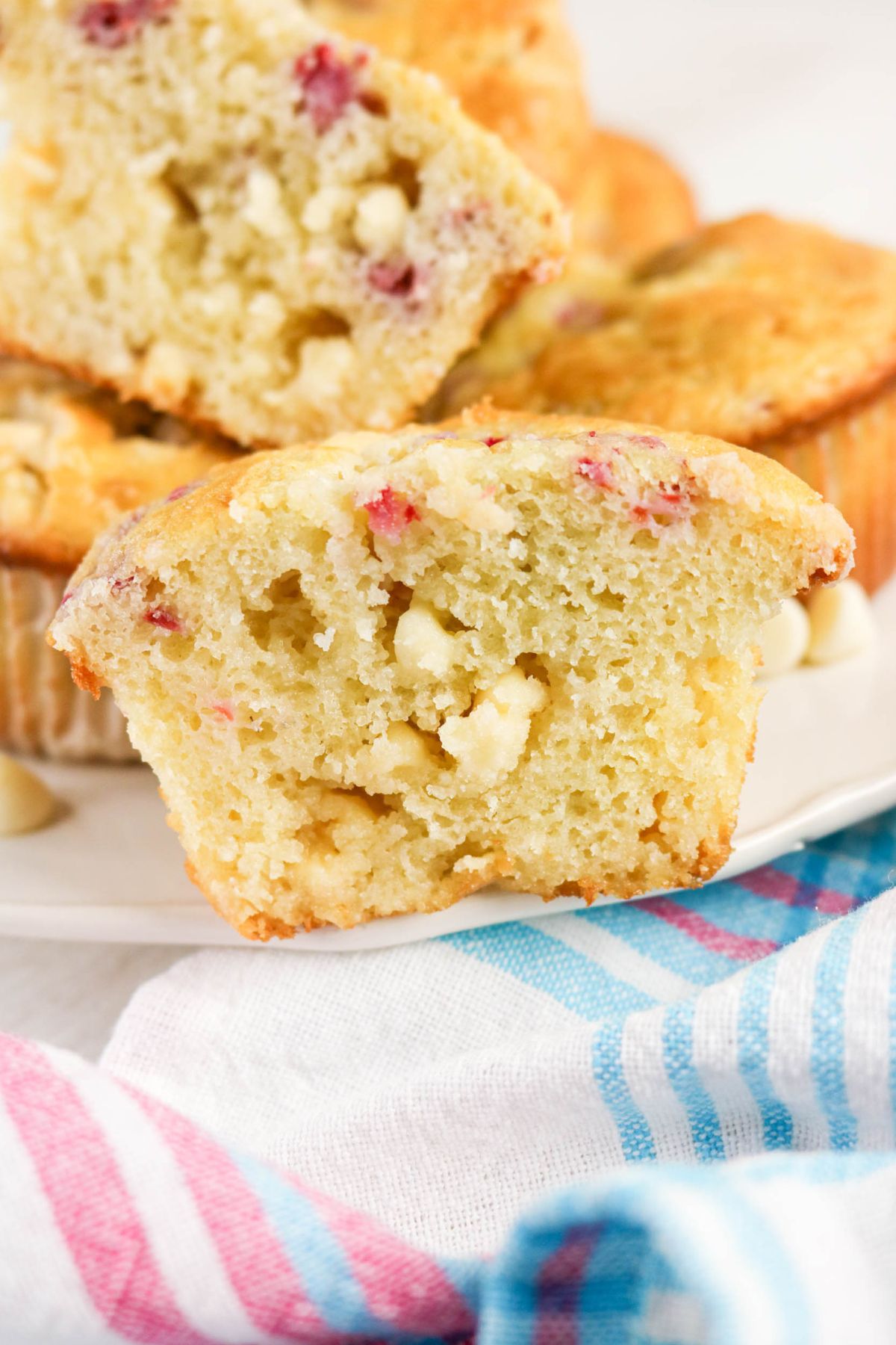 A close-up of a sliced muffin with visible white chocolate chips and small pieces of red fruit, resting on a white plate near a striped cloth.
