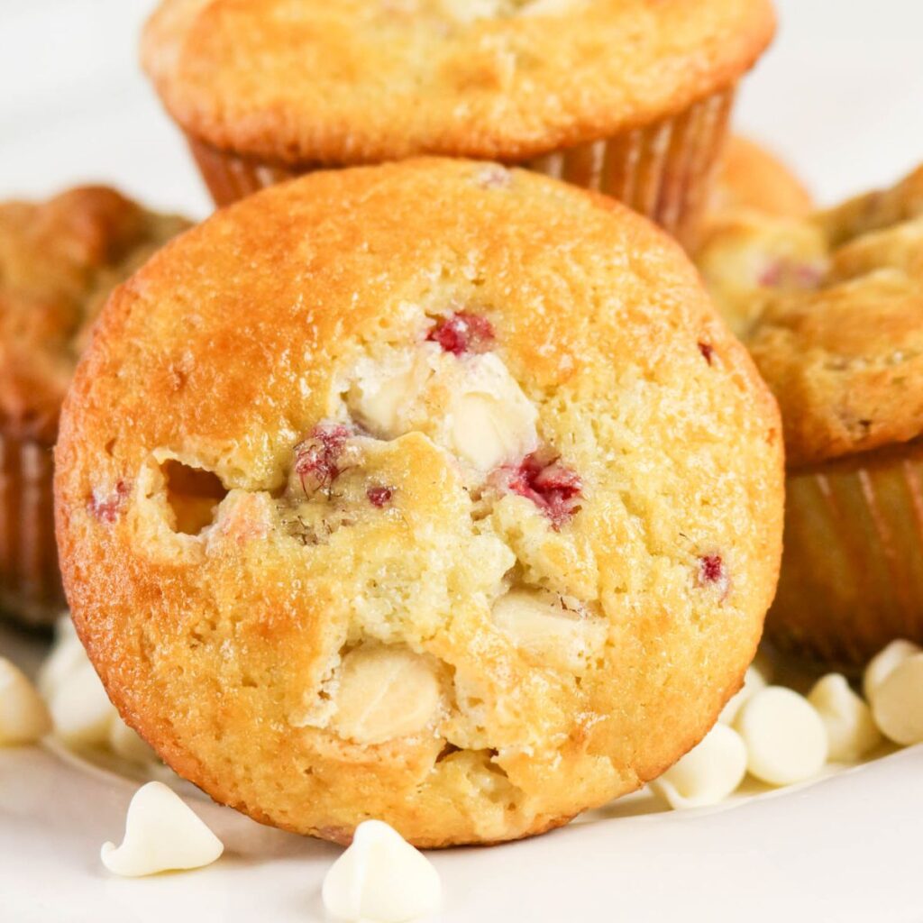 Close-up of a raspberry white chocolate muffin on a plate, with white chocolate chips scattered around and more muffins in the background.