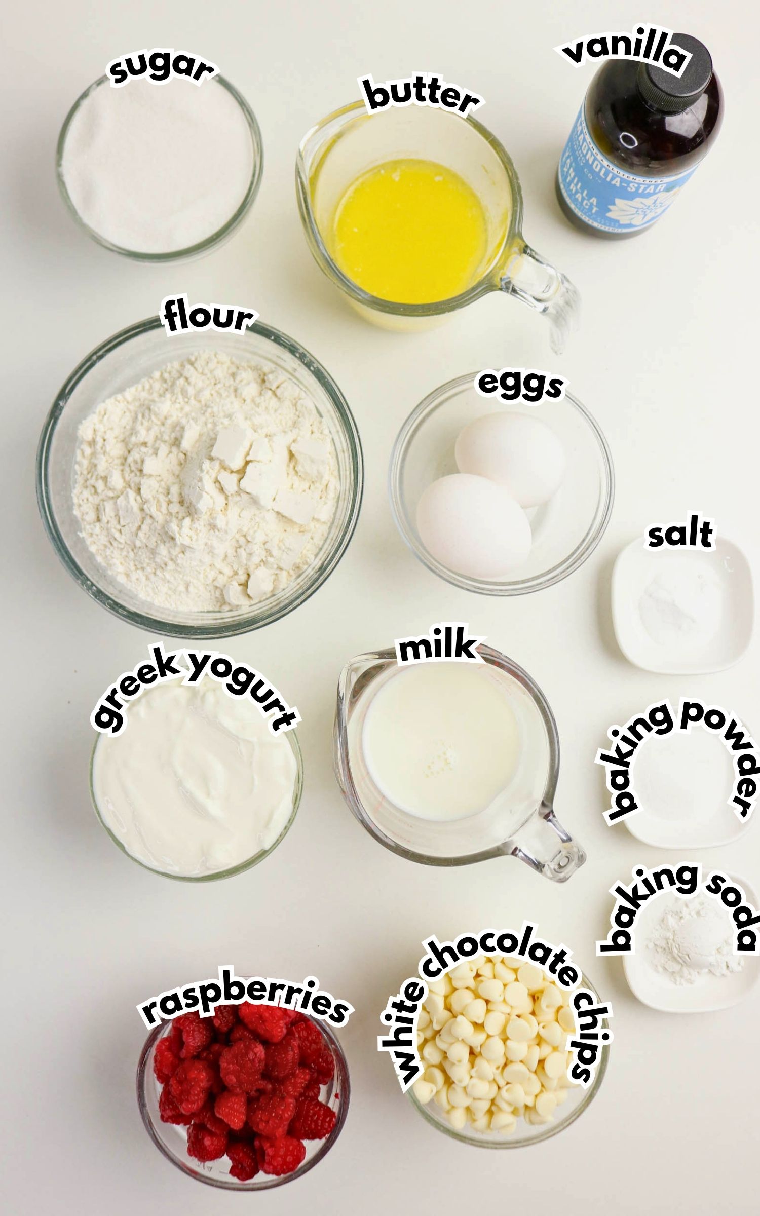 Top-down view of measured baking ingredients in bowls, including sugar, flour, butter, eggs, vanilla, Greek yogurt, milk, salt, baking powder, baking soda, raspberries, and white chocolate chips.