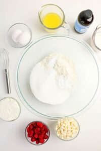 Various baking ingredients including flour, sugar, eggs, melted butter, vanilla extract, raspberries, white chocolate chips, yogurt, and milk are arranged around a mixing bowl and whisk on a white surface.
