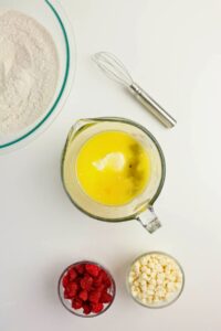 A mixing bowl with flour, a whisk, a measuring cup with wet ingredients, and small bowls of raspberries and white chocolate chips on a white surface.